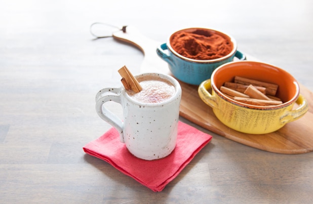 A mug of Mexican style hot chocolate beside bowls of chili powder and cinnamon sticks.