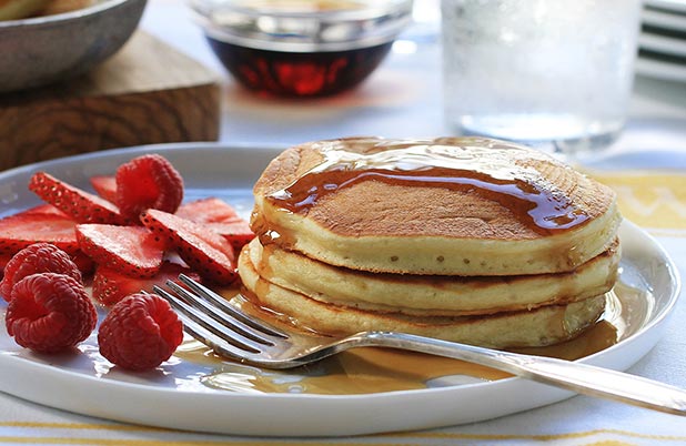 A stack of pancakes with syrup on a plate with a fork, strawberries and raspberries.