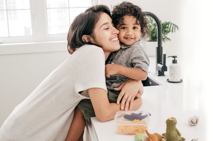 A young curly haired chil sits on the kitchen counter beside toys and snacks smiling while his young mom smiles and hugs him.