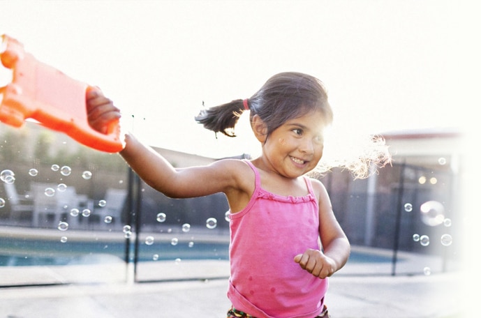A smiling girl in a bright pink shirt stands twirling a bubble maker around her leaving a trail of bubbles behind her .