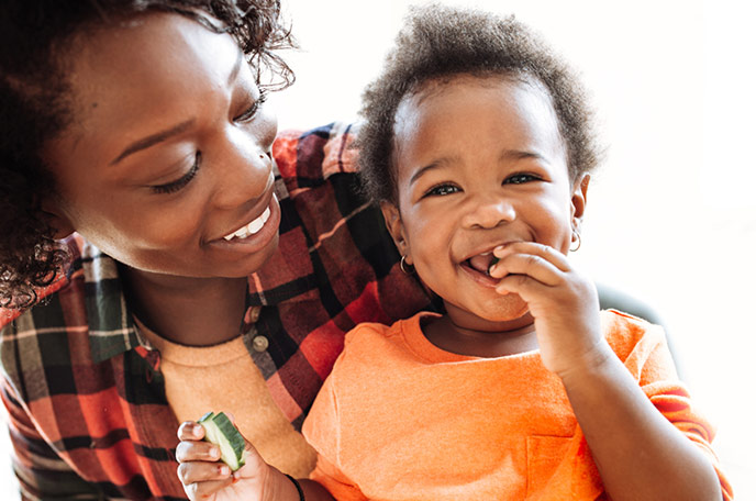 A young girl sits on her mothers lap and eats a cucumber slice.