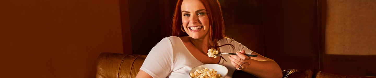 Woman sitting up on orange couch smiling to camera about to take a bite of Lean Cuisine Vermont White Cheddar Mac & Cheese.