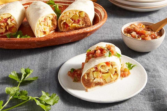 Breakfast burritos next to a bowl of salsa, plates, and cilantro on a textured grey surface.