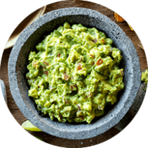 A stone mortar filled with guacamole surrounded by tortilla chips, lime wedges, cloth napkins, and avocado toast.