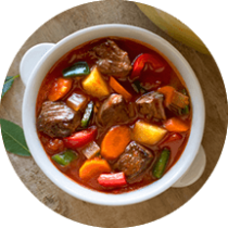 A white bowl of beef stew surrounded by bread, rice, bay leaves, a colorful pot and cloth napkin.