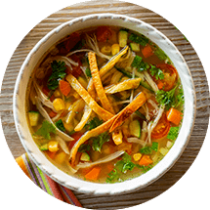 A bowl of tortilla soup with corn, red pepper, and tomato sits next to a striped cloth napkin.