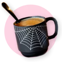 Un tarro decorado con una telaraña lleno de chocolate caliente y con una cuchara dentro sobre un círculo rosa.