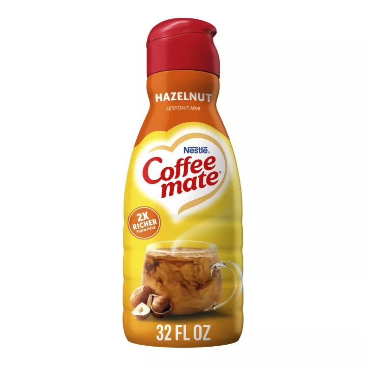 A bottle of Hazelnut Creamer with a red label above a coffee mug, whole hazelnuts and the product logo.
