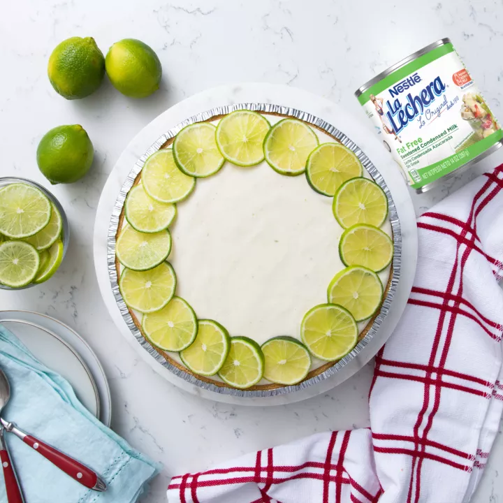 Una lata color blanco con las etiquetas color verde y el logo de La Lechera, junto a una copa de fruta rociada con leche condensada azucarada.
